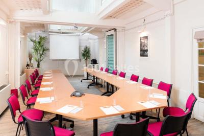 Salle de réunion avec table blanche, chaises rouges, écran mural et éclairage au plafond.