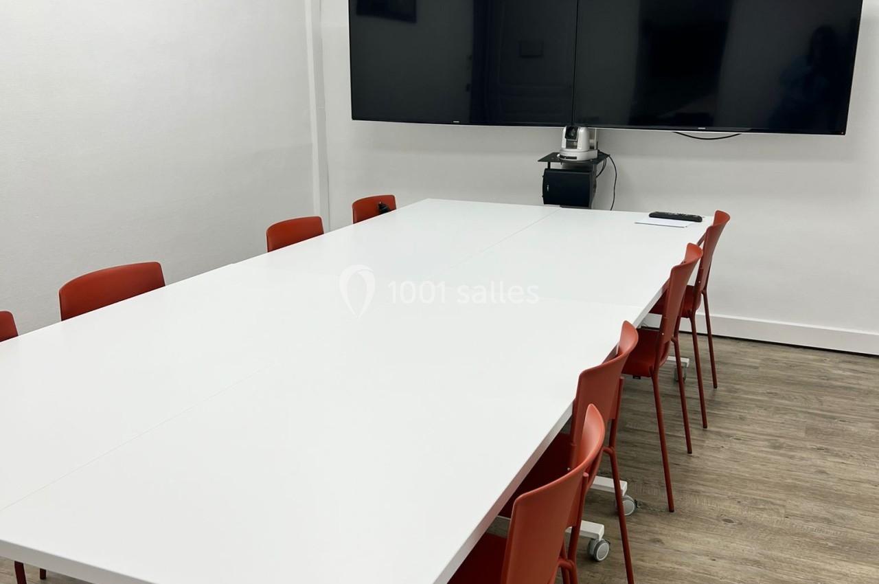 Salle de réunion avec une grande table blanche, des chaises rouges, deux écrans muraux et un sol en bois clair.