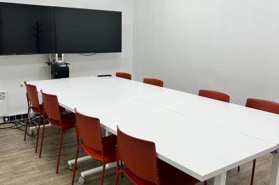 Salle de réunion avec table blanche, chaises rouges, écran mural et éclairage au plafond.