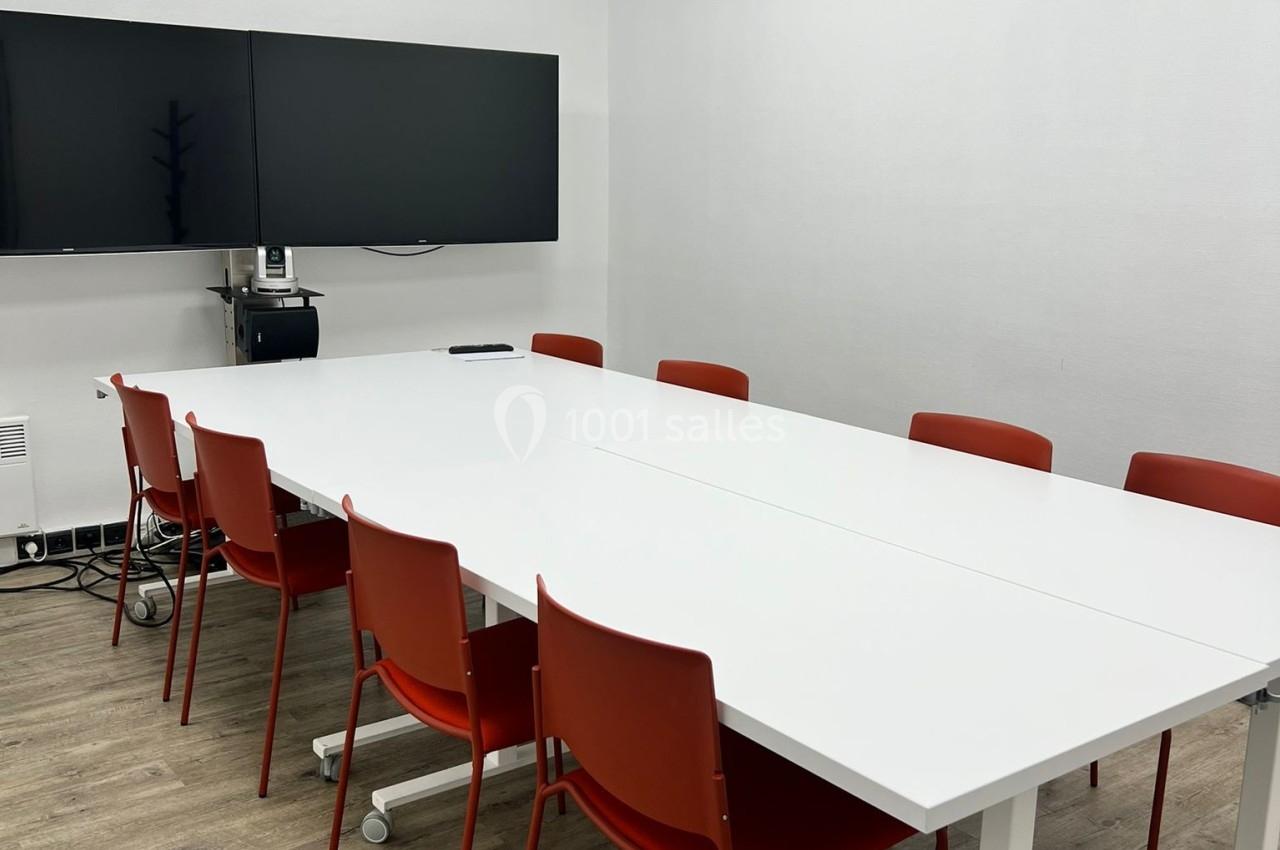 Salle de réunion avec table blanche, chaises rouges, deux écrans muraux et sol en parquet clair.