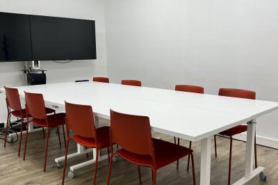 Salle de réunion avec table blanche, chaises rouges, écran mural et éclairage au plafond.