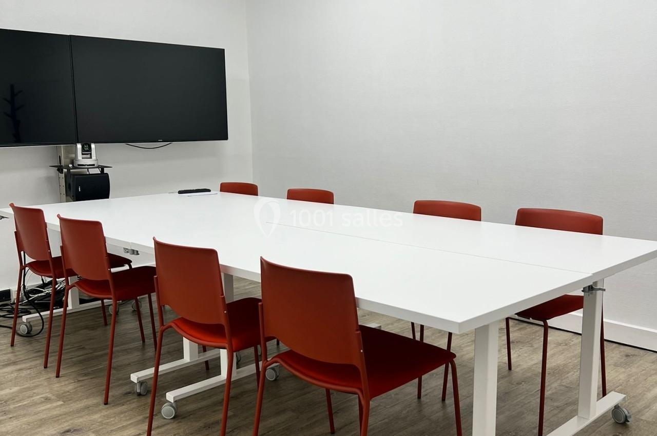 Salle de réunion avec table blanche, chaises rouges, écran mural et sol en parquet clair.