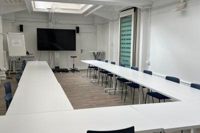 Salle de réunion avec table blanche, chaises rouges, écran mural et éclairage au plafond.
