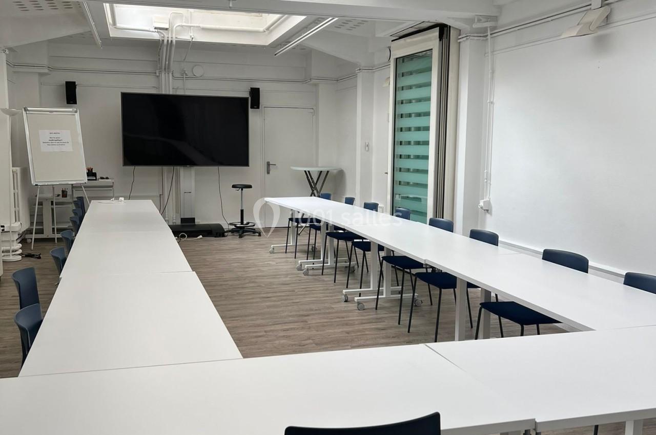 Salle de réunion lumineuse avec tables blanches en disposition en U, chaises noires et écran de présentation.