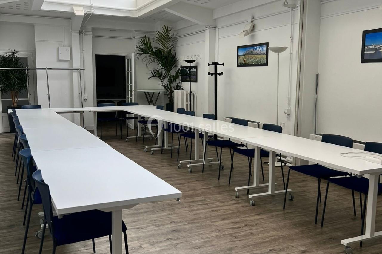 Salle de réunion lumineuse avec de grandes tables blanches disposées en U, chaises bleues et plantes en arrière-plan.