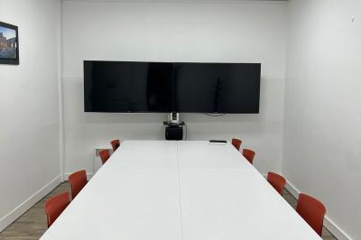 Salle de réunion avec table blanche, chaises rouges, écran mural et éclairage au plafond.