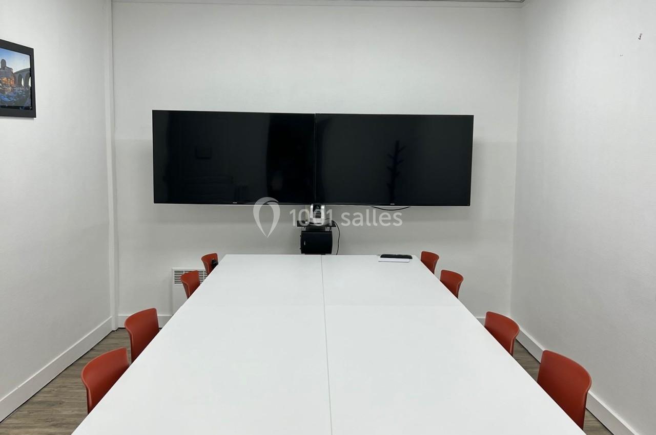 Salle de réunion avec table blanche, chaises rouges, écran mural et éclairage au plafond.