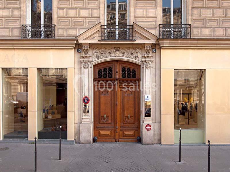 Façade d'un immeuble avec une porte en bois sculptée, encadrée par deux vitrines et des potelets sur le trottoir.