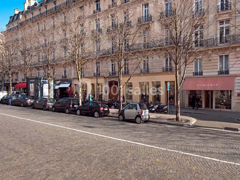 Façade d'immeubles haussmanniens avec commerces au rez-de-chaussée, arbres et voitures stationnées le long de la rue.