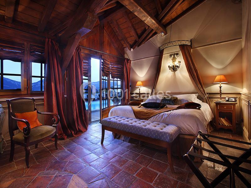 Chambre chaleureuse avec lit double, poutres apparentes, éclairage tamisé et vue sur des montagnes au crépuscule.