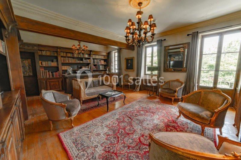 Salon chaleureux avec parquet, tapis rouge, bibliothèque en bois, fauteuils anciens et grandes fenêtres donnant sur l…