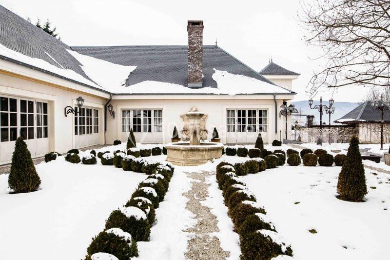 Cour enneigée avec une fontaine centrale entourée de buis taillés, devant une maison aux toits d'ardoise.