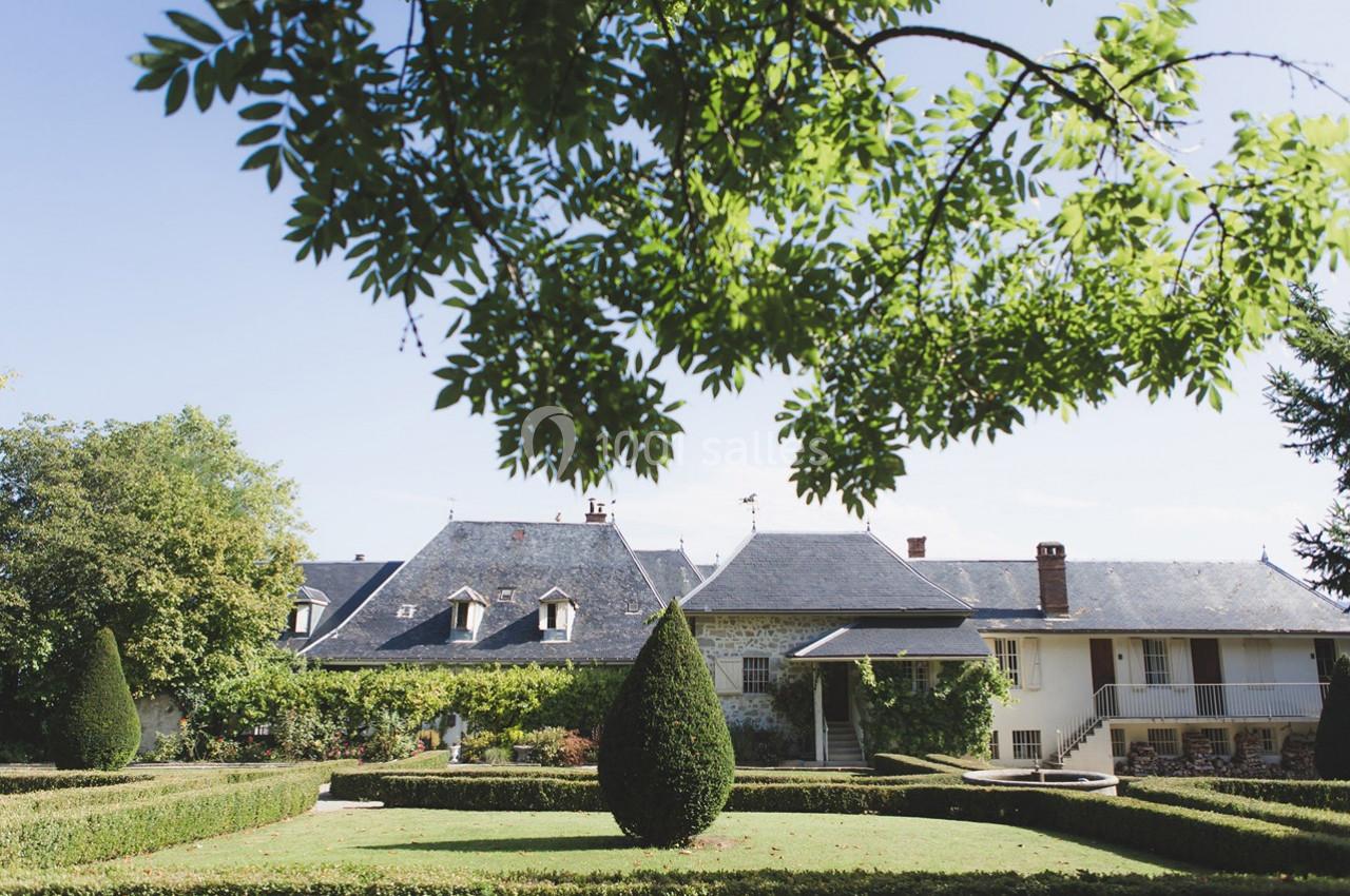 Jardin avec haies taillées et arbres, devant une maison traditionnelle aux toits en ardoise sous un ciel dégagé.