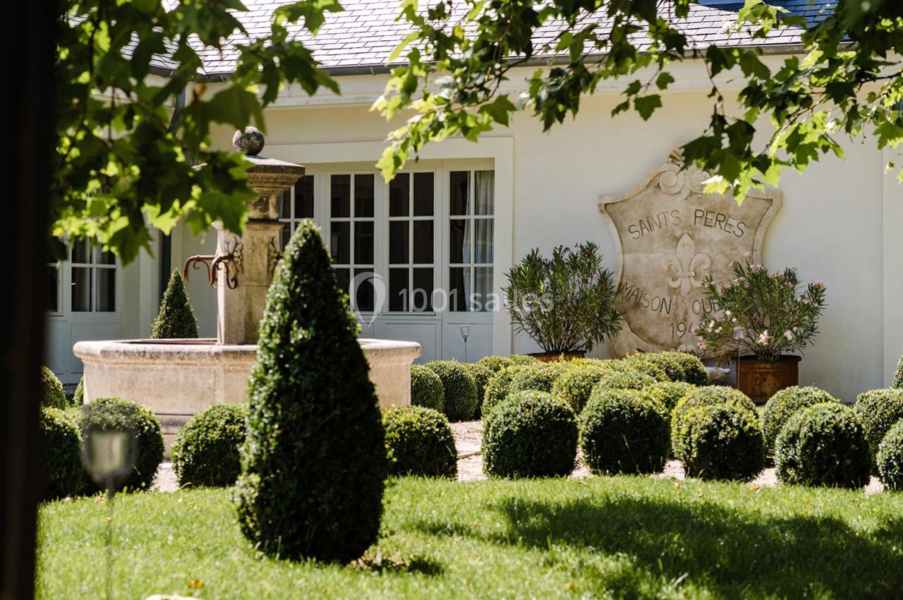 Jardin avec fontaine centrale, buis taillés et façade blanche ornée d'une plaque gravée.
