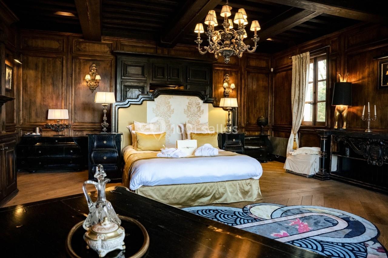 Chambre élégante avec lit double, boiseries sombres, lustre, tapis décoratif et lumière naturelle entrant par une fenêtre.