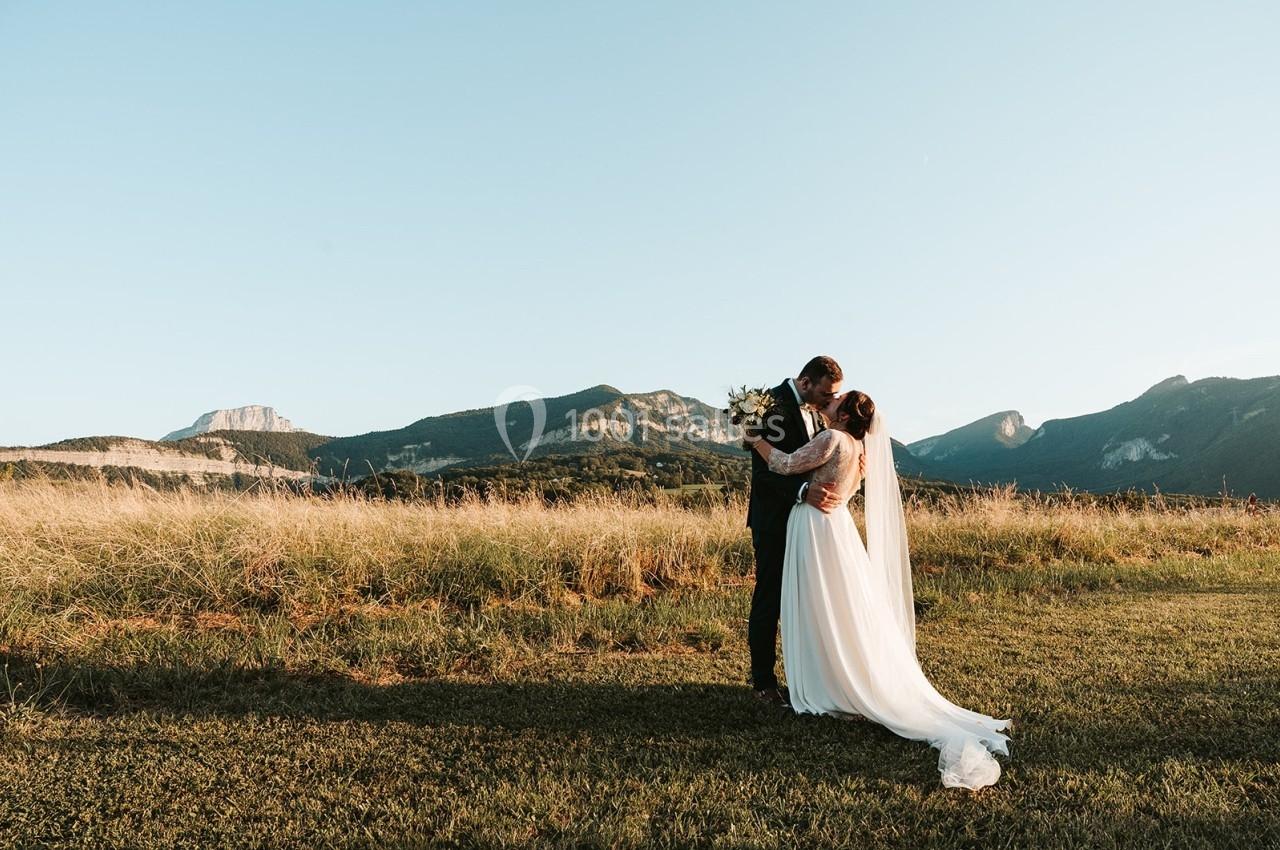Un couple en tenue de mariage s'embrasse dans un champ au coucher du soleil, avec des montagnes en arrière-plan.