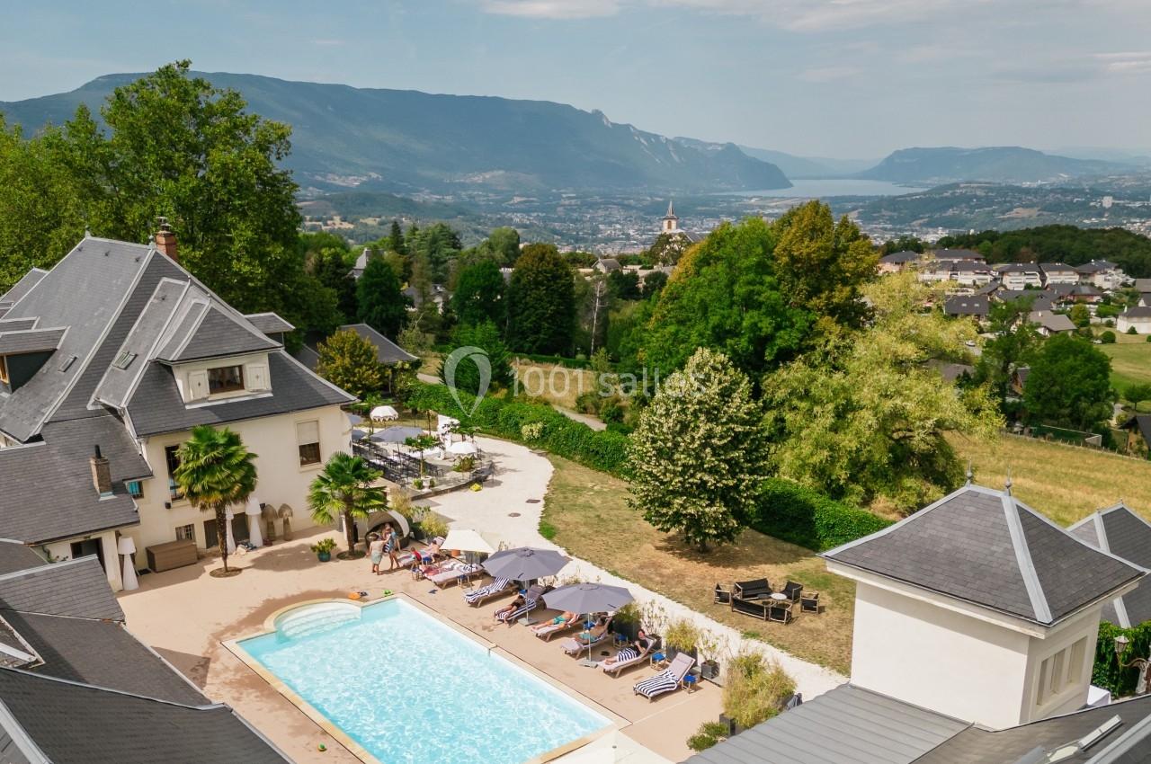 Vue aérienne d'une maison avec piscine entourée de verdure, offrant un panorama sur des montagnes et un lac au loin.