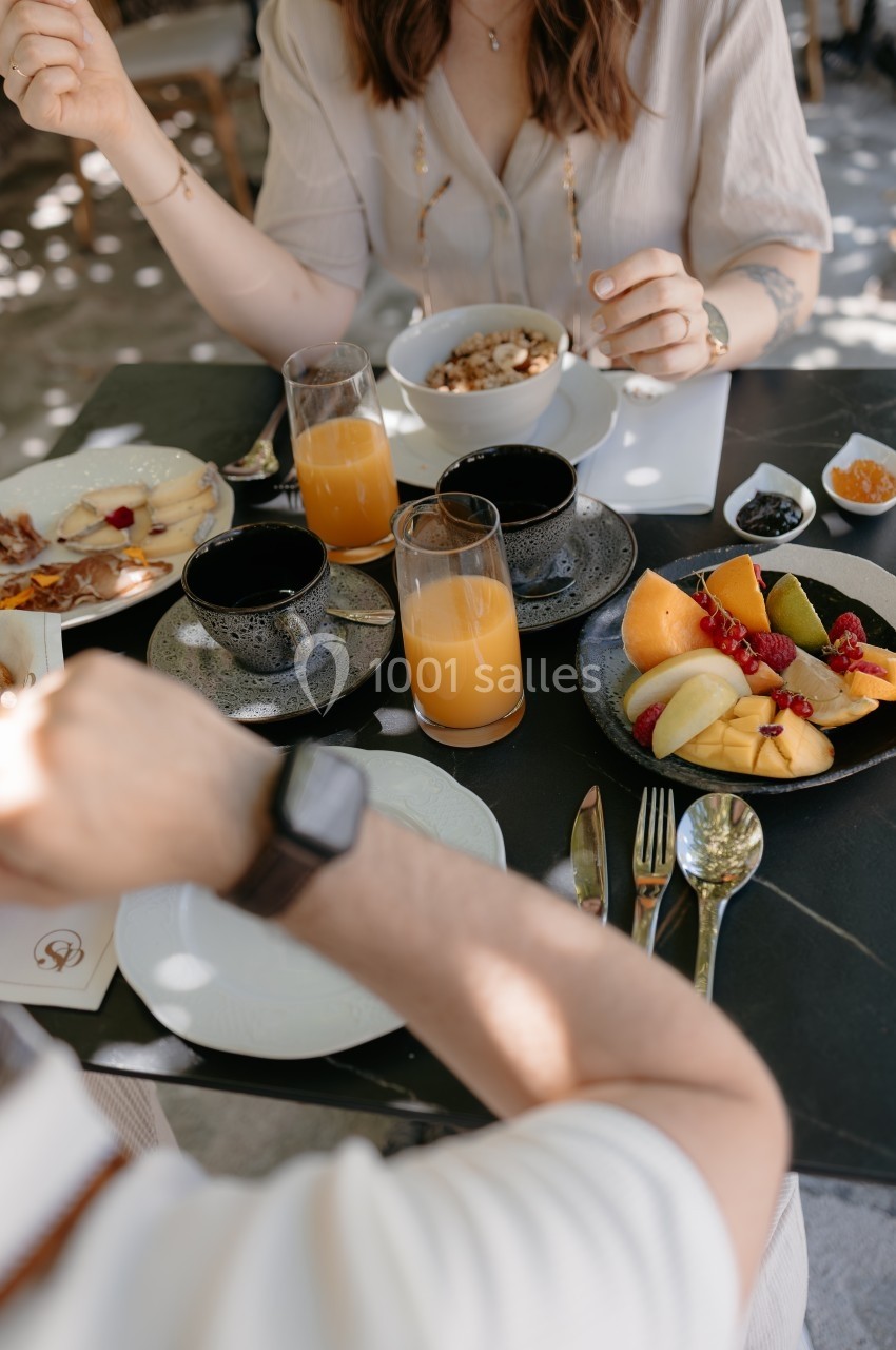 Deux personnes prennent un petit-déjeuner avec fruits, jus d'orange, céréales et couverts sur une table noire.