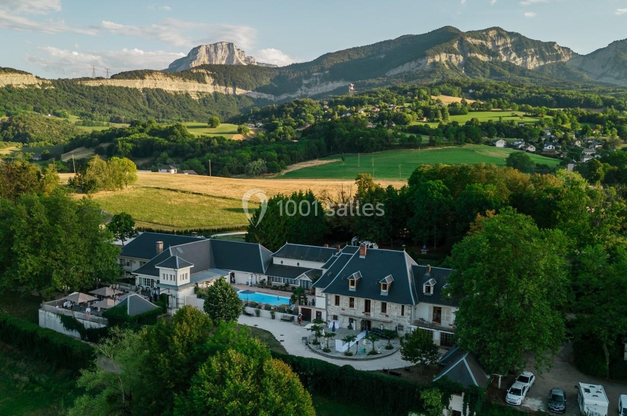 Vue aérienne d'un domaine entouré de verdure avec des montagnes en arrière-plan sous un ciel partiellement nuageux.