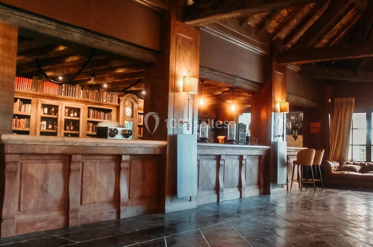 Intérieur d'un bar chaleureux avec comptoir en bois, étagères de livres, éclairage tamisé et fauteuils près d'une fenêtre.