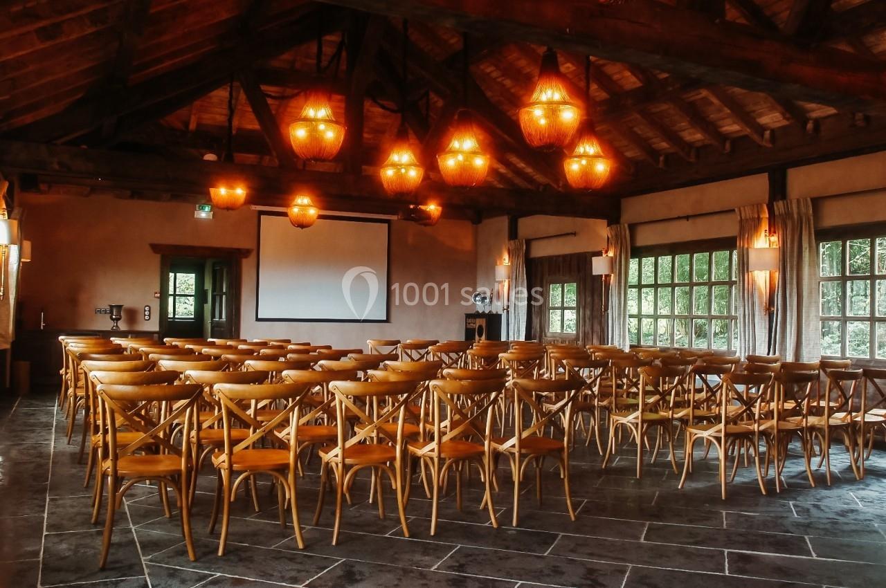 Salle de conférence avec des chaises en bois disposées face à un écran de projection, éclairée par des lustres suspendus.