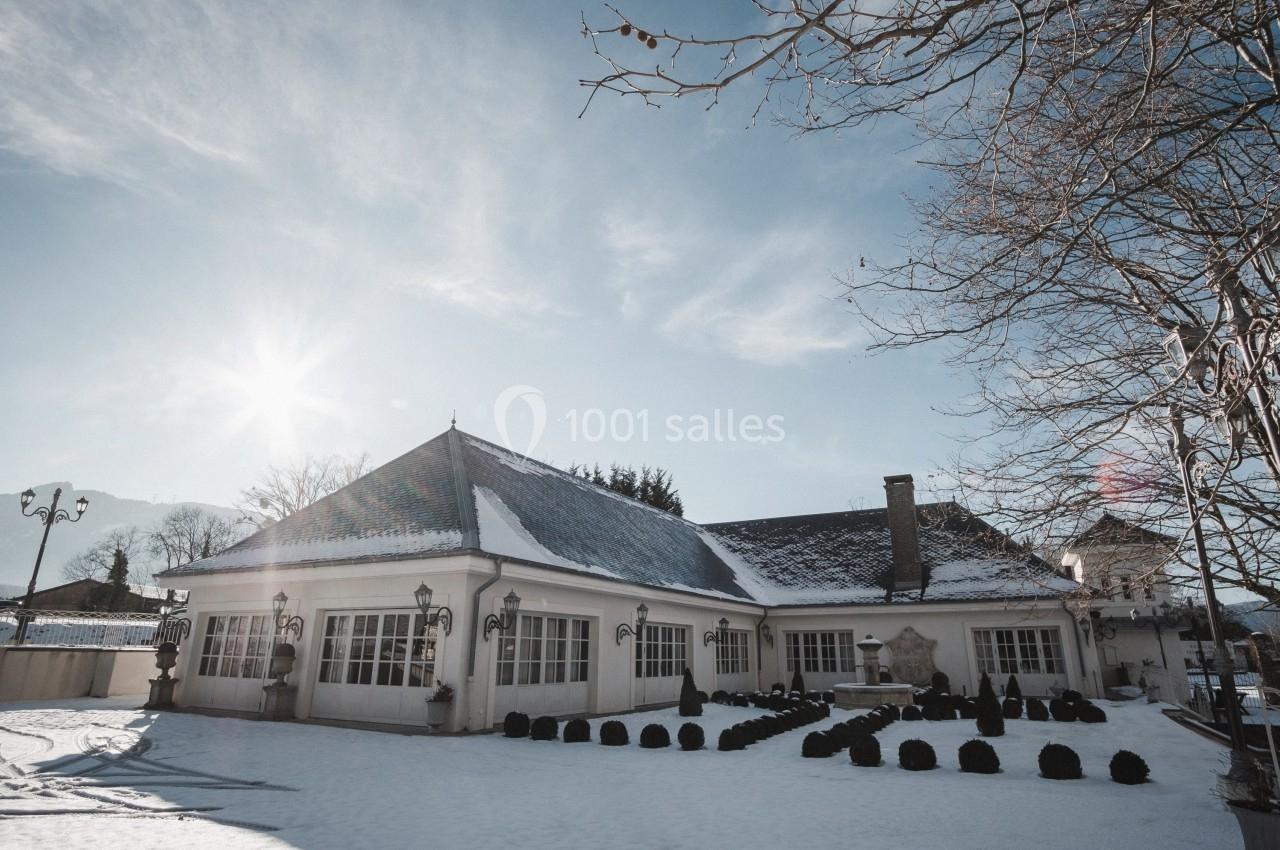 Bâtiment avec toit enneigé, grandes fenêtres et éclairage extérieur, entouré d'un jardin sous un ciel ensoleillé.