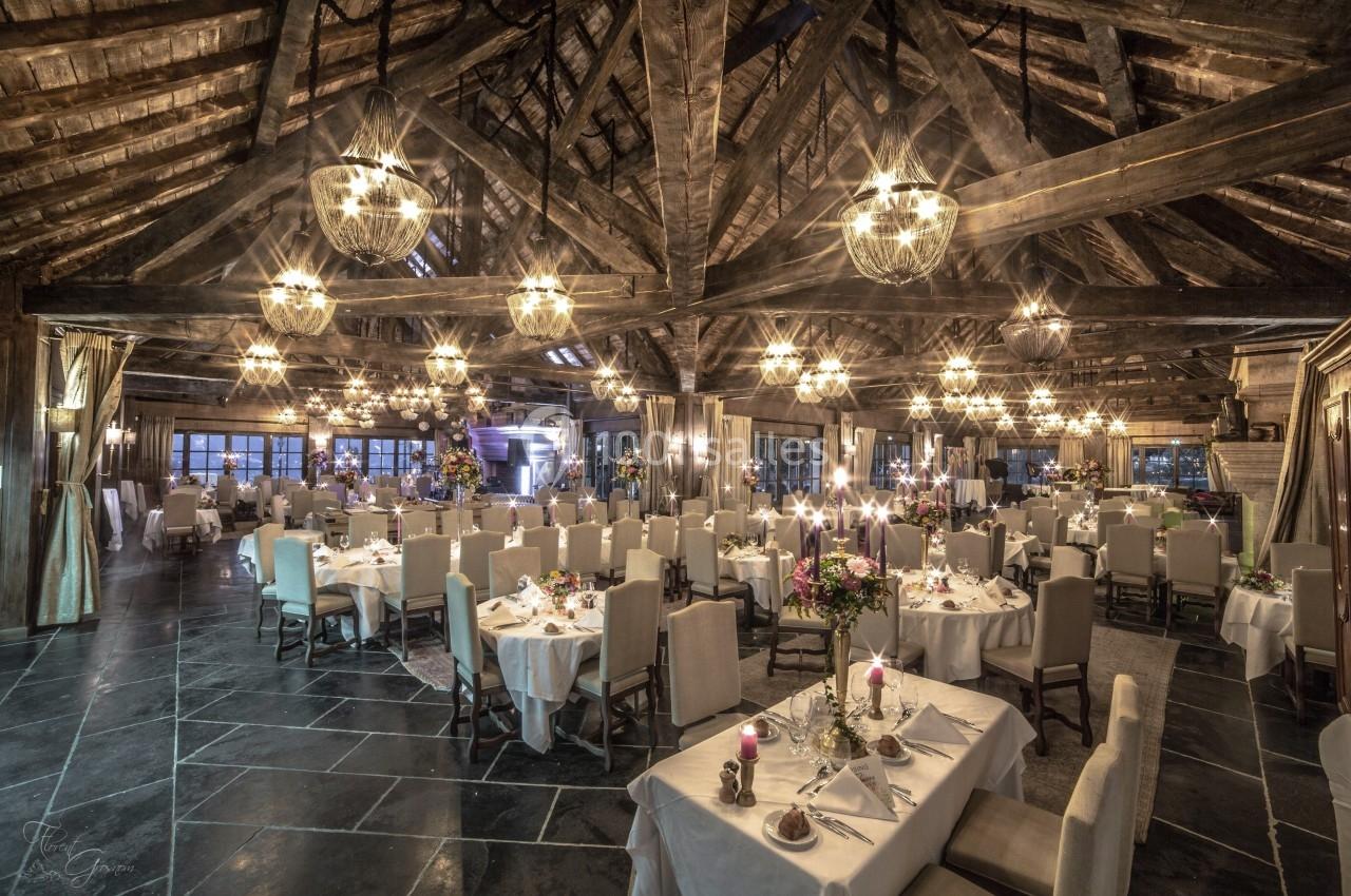 Salle de réception élégante avec tables dressées, chandeliers allumés et poutres apparentes en bois.