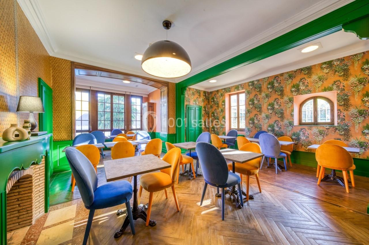 Salle lumineuse avec tables et chaises colorées, parquet en bois, murs décorés de papier peint floral et cheminée.