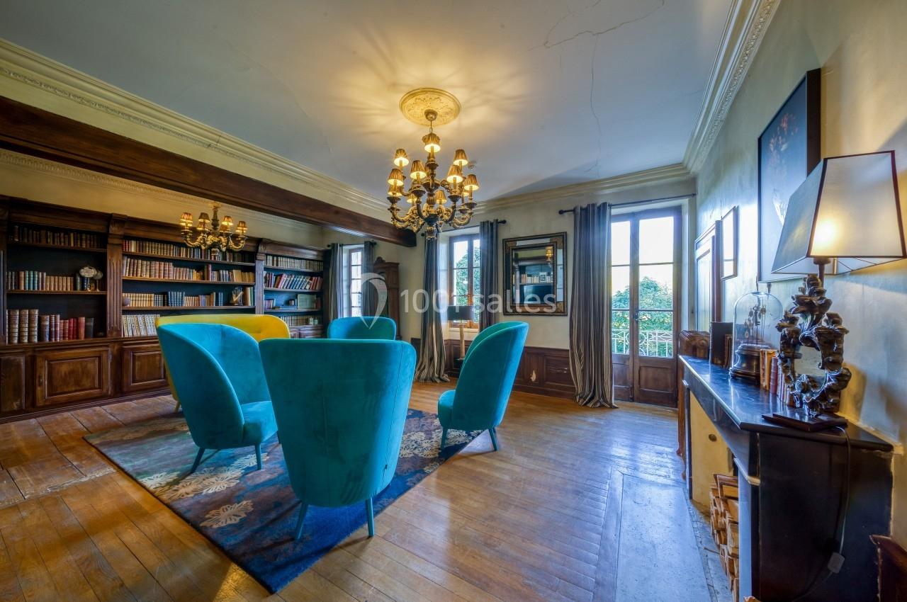 Salon lumineux avec fauteuils bleus, bibliothèque en bois, lustres anciens et grandes fenêtres donnant sur un jardin.