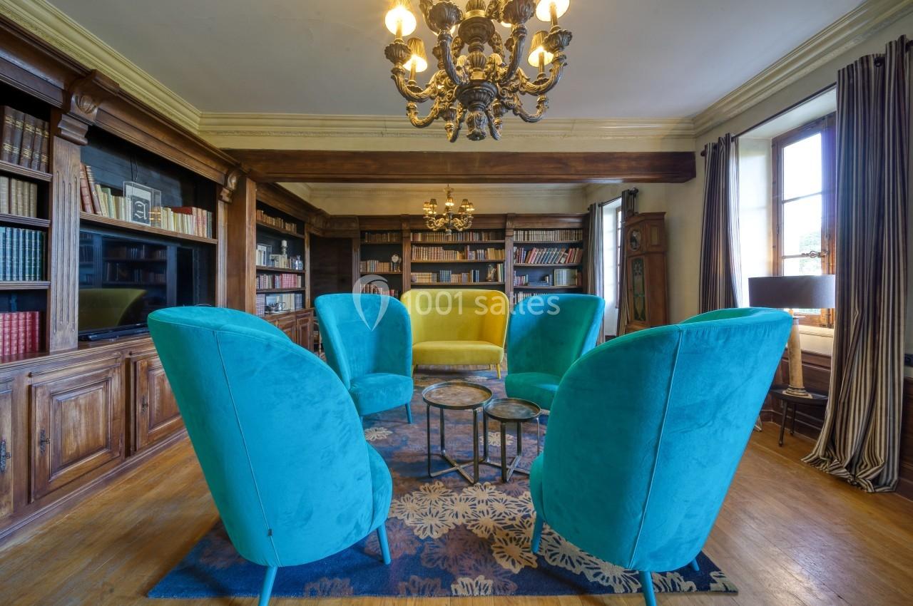 Salon avec fauteuils turquoise et jaune disposés en cercle, bibliothèque en bois, lustres et grande fenêtre en arrière-plan.