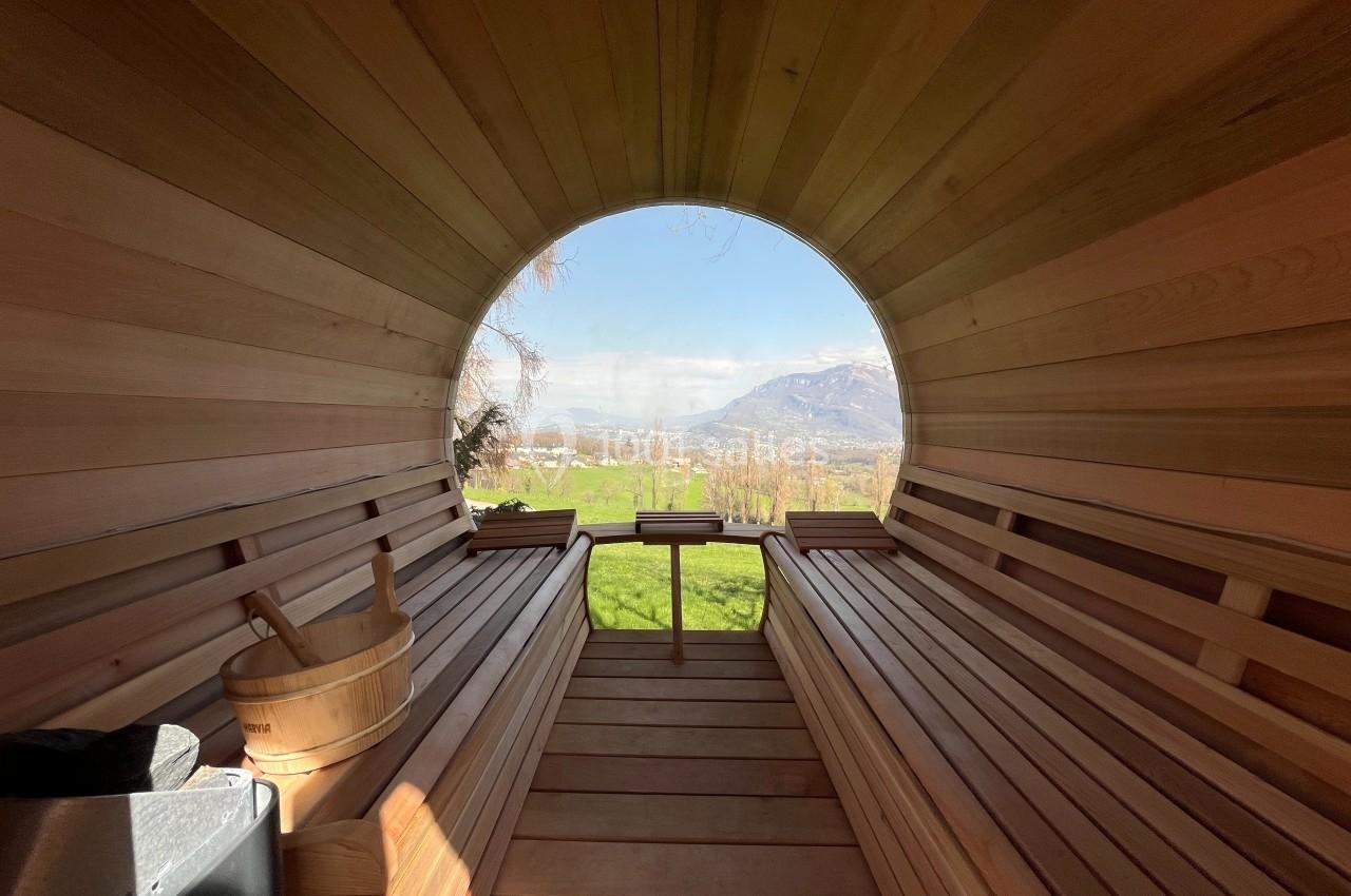Intérieur d'un sauna en bois en forme de tonneau avec vue sur un paysage verdoyant et des montagnes au loin.