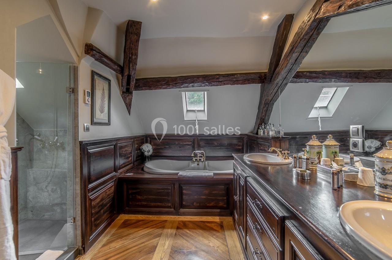 Salle de bain sous combles avec poutres apparentes, baignoire encastrée, double vasque et parquet en bois.