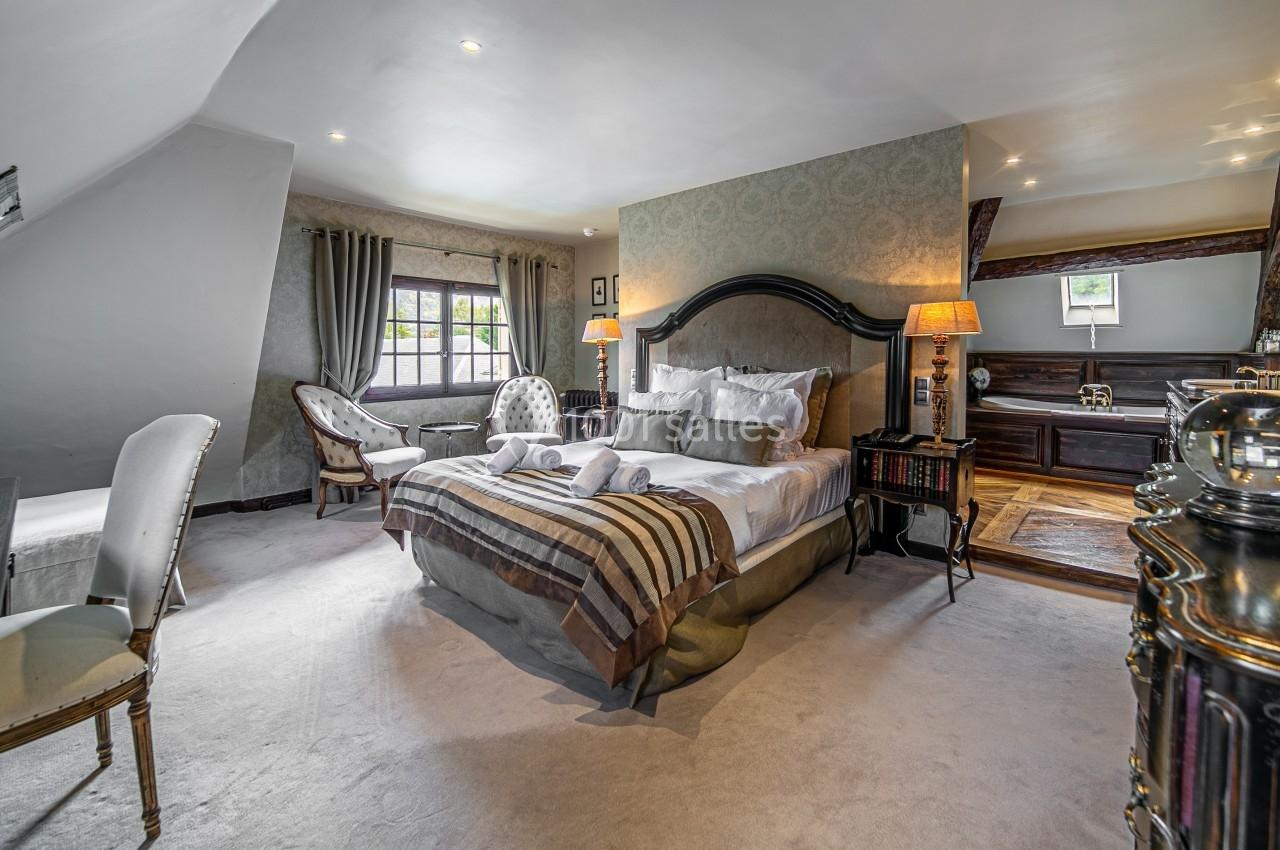 Chambre élégante avec lit double, fauteuils près d'une fenêtre, mobilier en bois sombre et éclairage chaleureux.