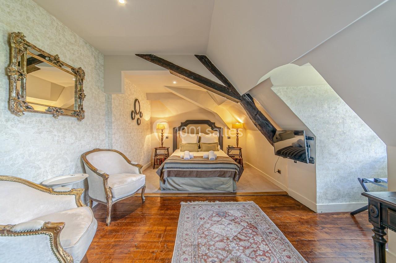 Chambre mansardée avec lit double, poutres apparentes, fauteuils anciens, miroir doré et tapis sur parquet.