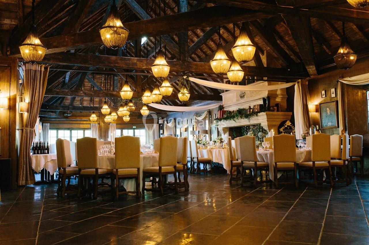 Salle de réception rustique avec poutres apparentes, tables dressées et éclairage chaleureux par des lustres suspendus.