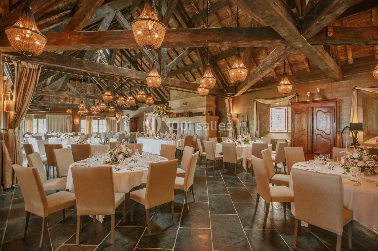 Salle de réception avec tables rondes dressées, chaises beige clair et poutres apparentes sous un éclairage chaleureux.