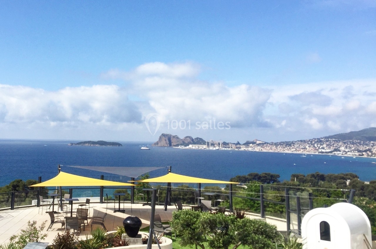 Vue sur une terrasse avec des voiles d'ombrage jaunes, donnant sur la mer et une ville côtière au loin.