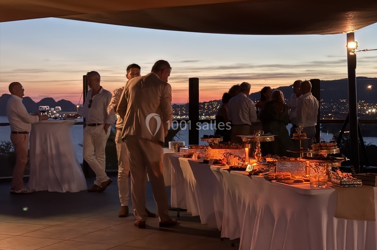 Groupe de personnes discutant près d'un buffet éclairé au coucher du soleil, avec vue sur une ville et la mer.