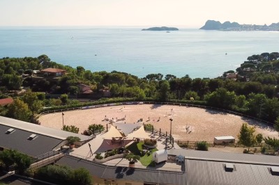 Vue aérienne d'un centre équestre entouré de végétation, avec la mer et des îles en arrière-plan.