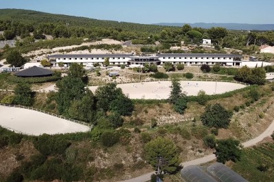 Vue aérienne d'un centre équestre entouré de végétation, avec la mer et des îles en arrière-plan.