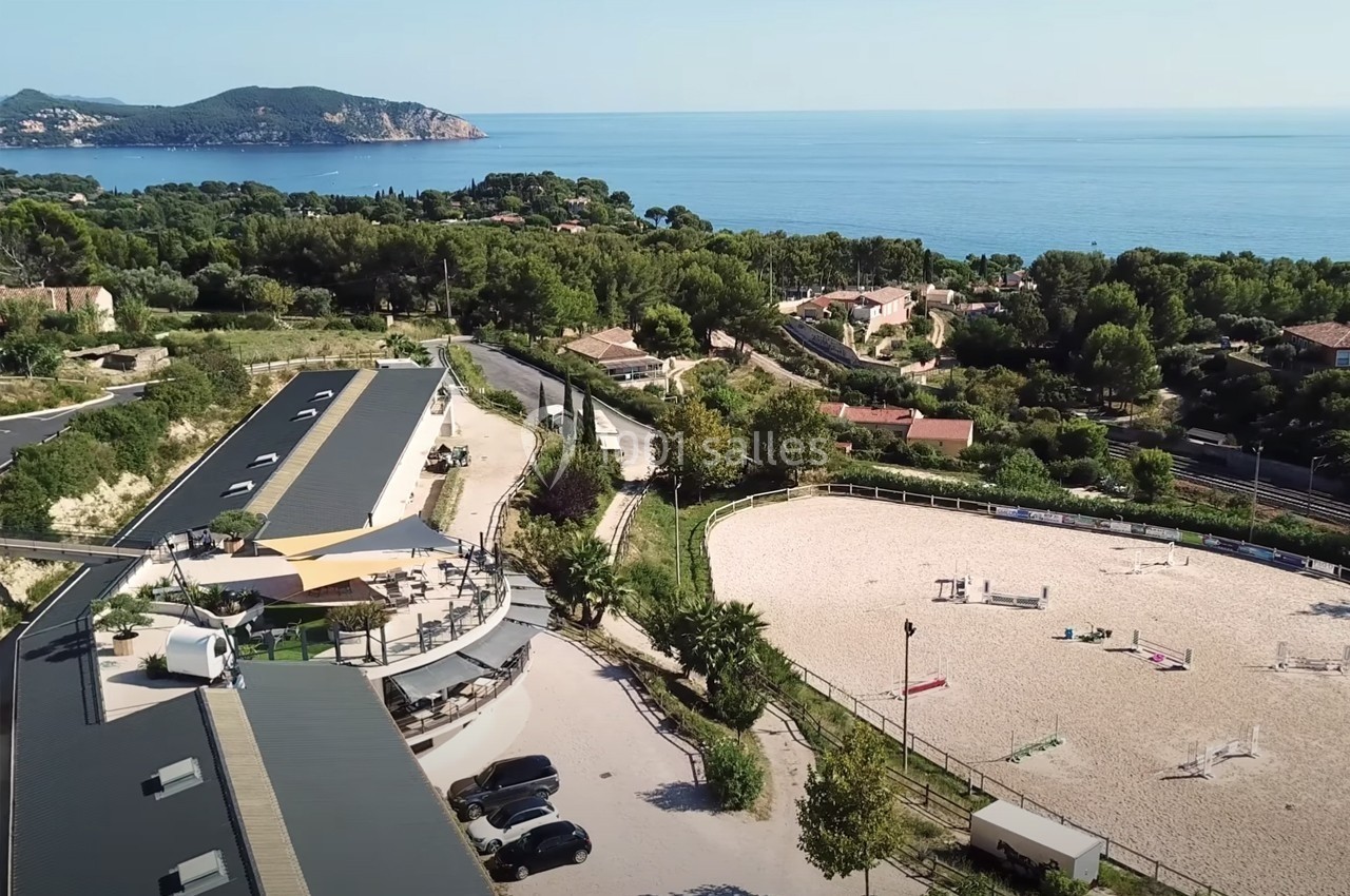 Vue aérienne d'un centre équestre avec carrière de sable, bâtiments, végétation et mer en arrière-plan.