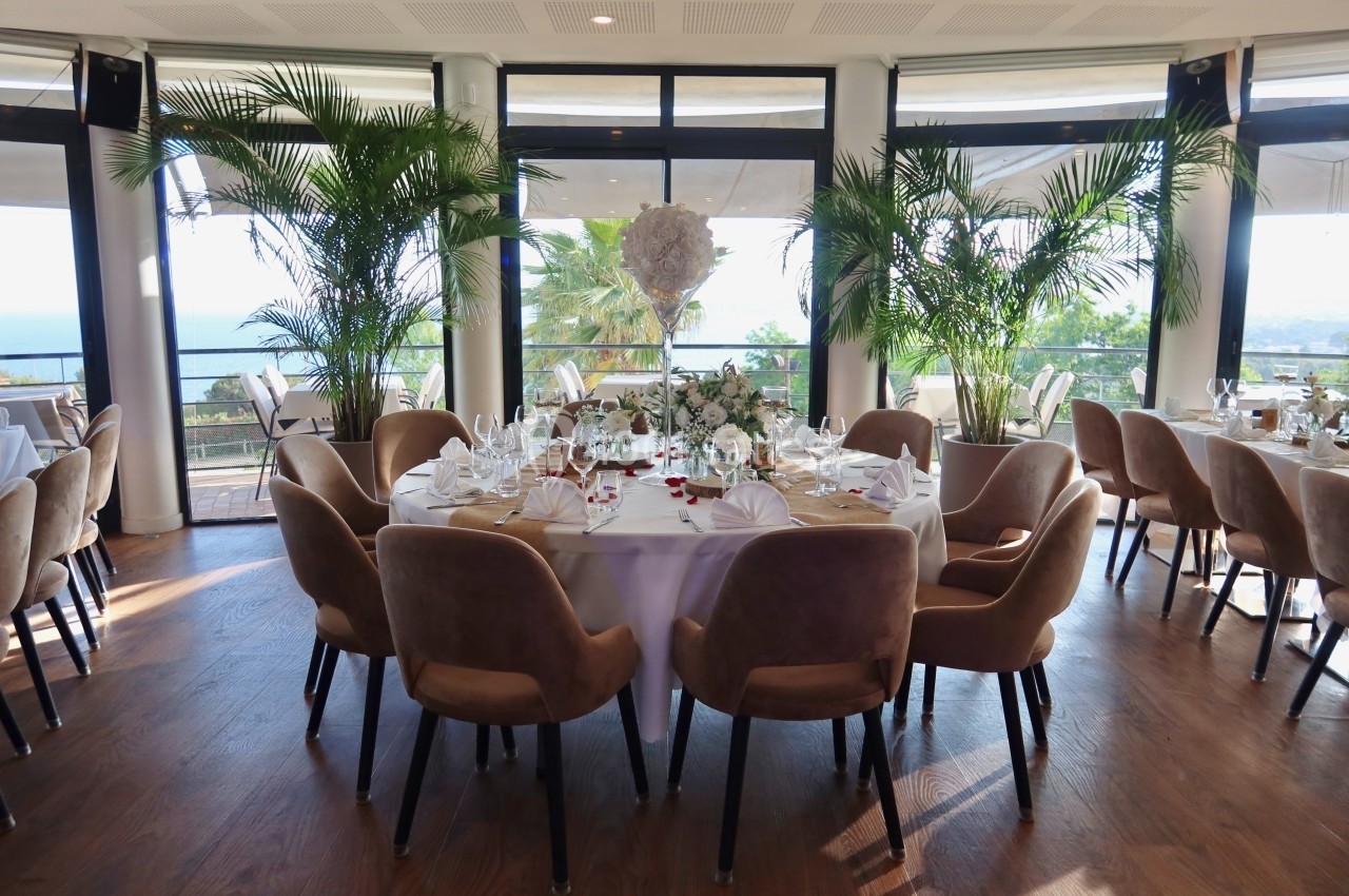 Salle de réception lumineuse avec tables rondes décorées, chaises en velours beige et vue sur la mer à travers de grandes…