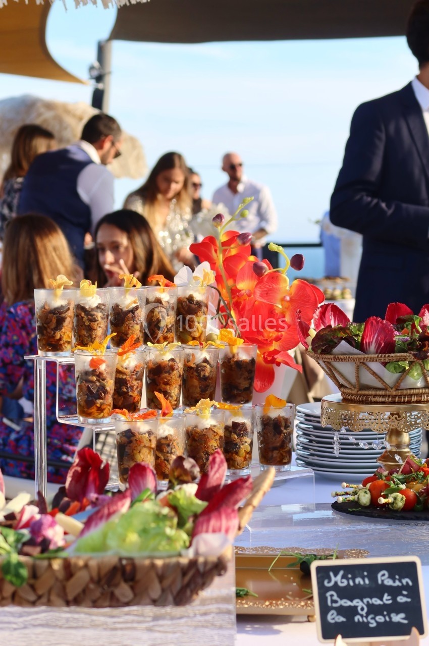 Buffet en plein air avec verrines, fleurs décoratives rouges et ardoise indiquant ’Mini Pain Bagnat à la Niçoise’.