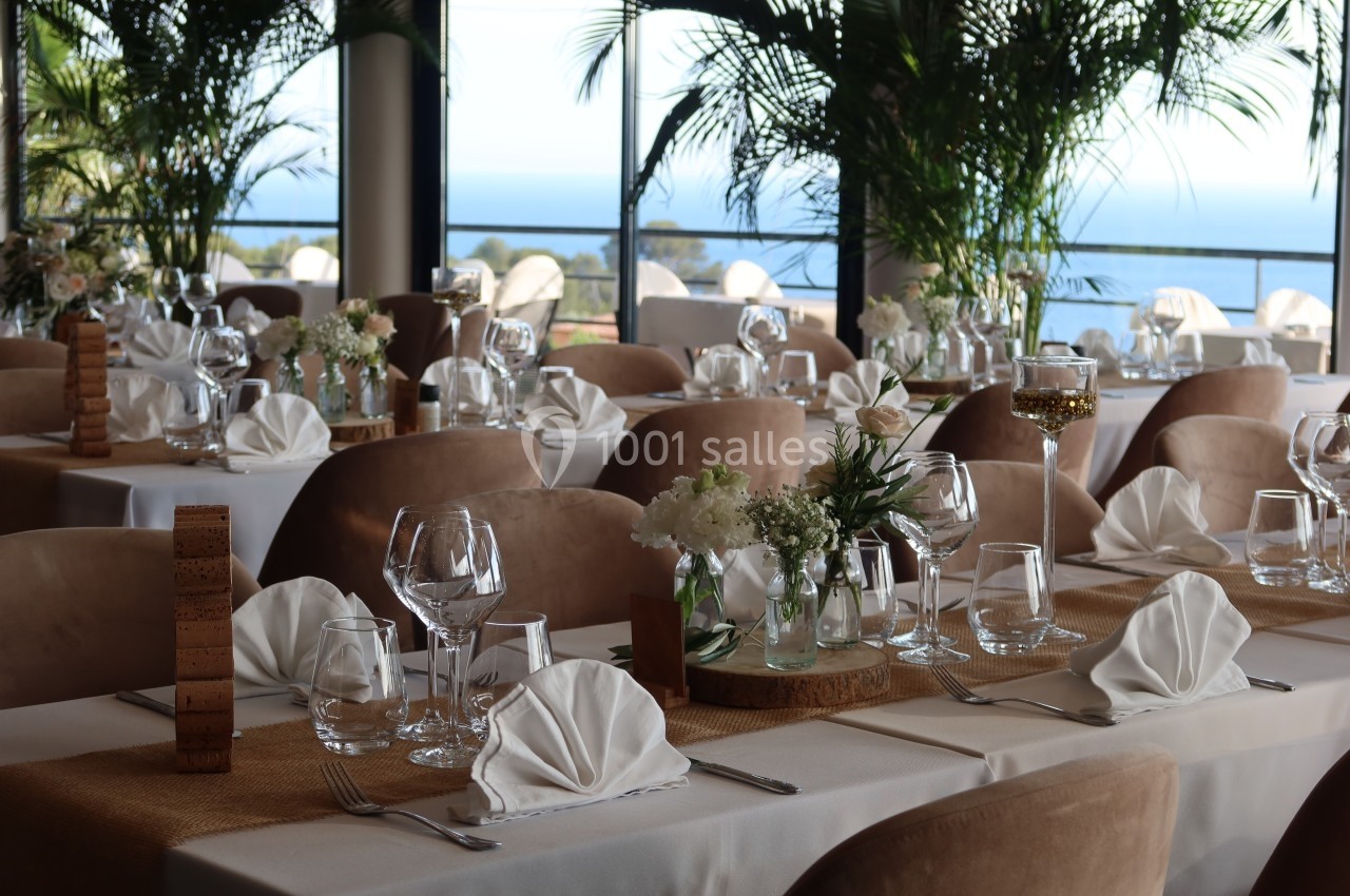 Salle de réception élégante avec tables dressées, nappes blanches, verres, serviettes pliées et vue sur la mer.