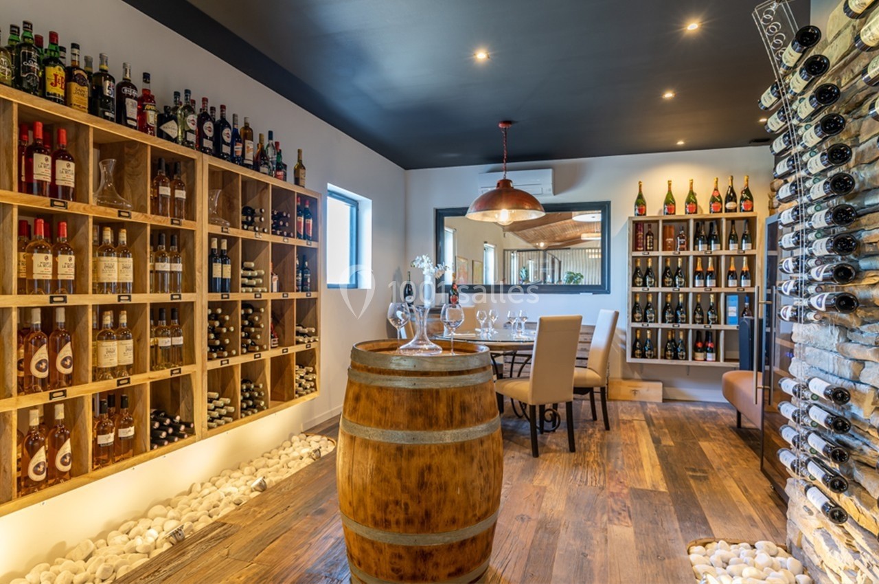 Salle de dégustation avec barriques en bois, étagères de bouteilles de vin et table dressée sous un éclairage chaleureux.