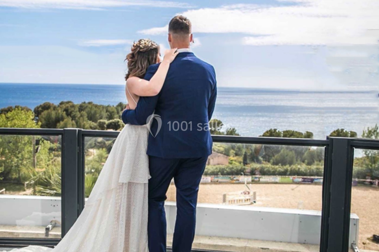 Un couple en tenue de mariage regarde la mer depuis une terrasse avec une vue dégagée sur un paysage côtier.
