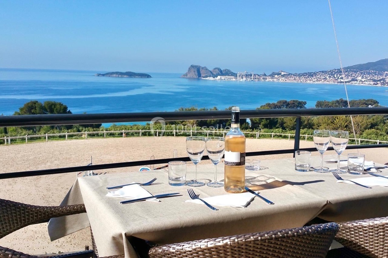 Table dressée avec une bouteille de vin et des verres, offrant une vue panoramique sur la mer et une côte ensoleillée.