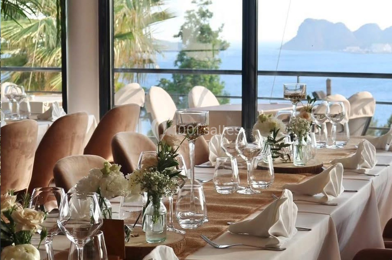 Salle de restaurant élégante avec vue sur la mer, tables dressées avec nappes blanches, fleurs et verrerie.