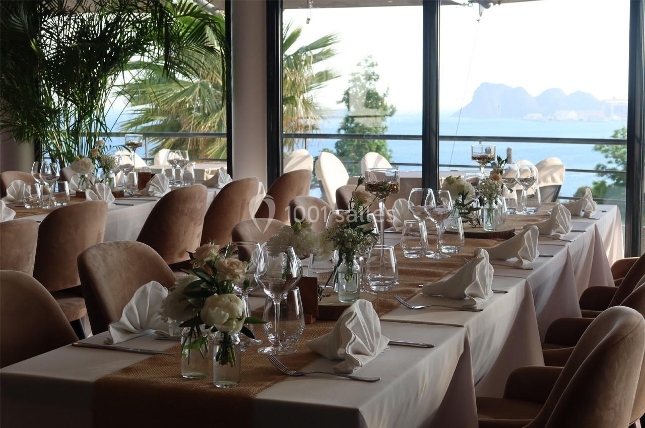 Salle de réception élégante avec tables dressées, nappes blanches, décor floral et vue sur la mer en arrière-plan.