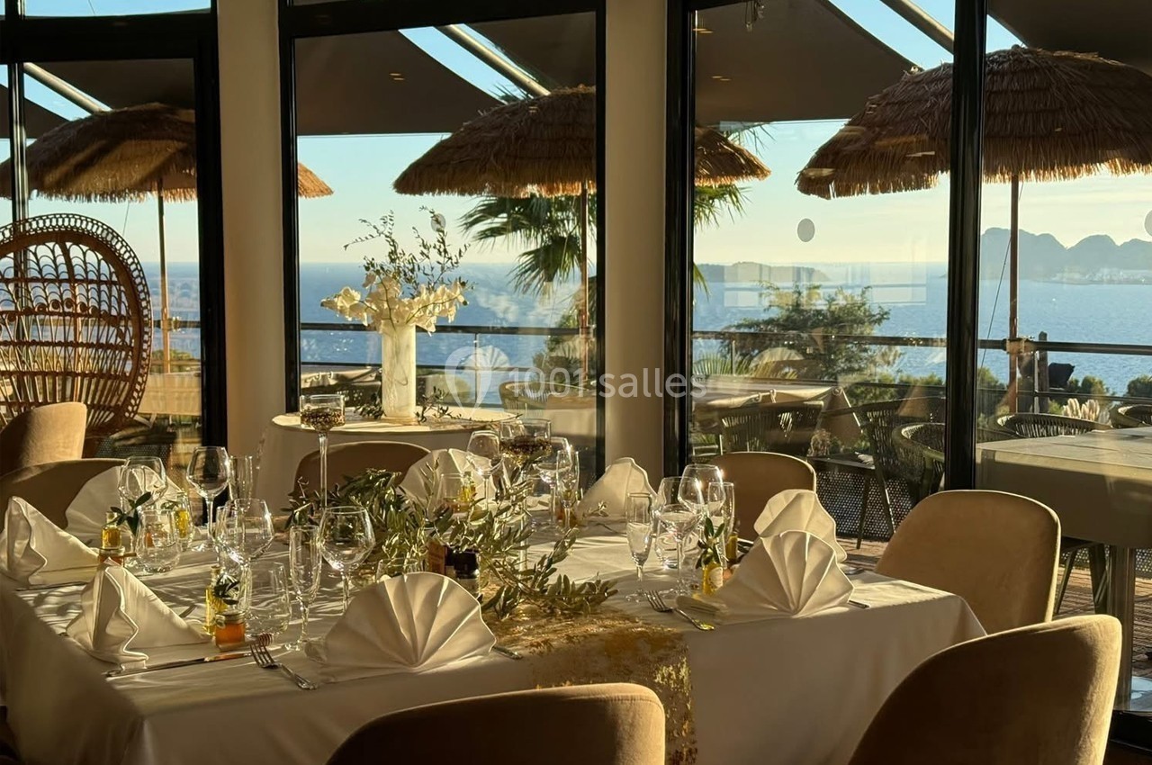 Salle de restaurant élégante avec vue sur la mer, tables dressées avec nappes blanches, décorations florales et lumière…